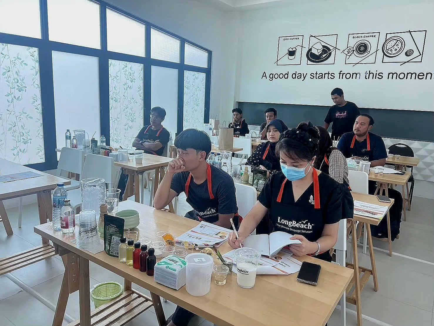 ผู้เข้าร่วมสวมผ้ากันเปื้อนนั่งประจำโต๊ะพร้อมวัตถุดิบเครื่องดื่มในห้องสัมมนา